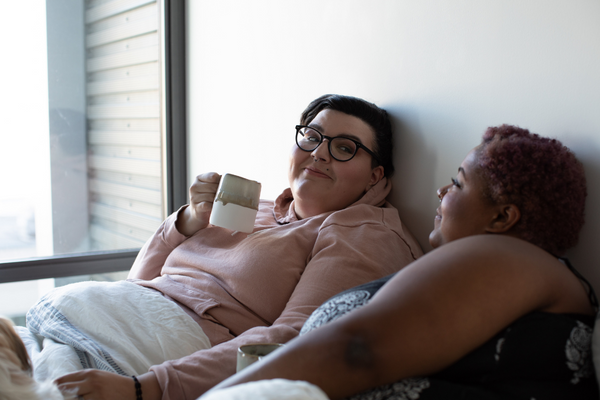 Two fat women lie in bed together and lean against the white wall behind them. The white woman on the left has short, brown hair, wears glasses and pink hoodie, and holds a white and tan coffee cup. The Black woman on the right has short, curly red hair. She wears a navy blue tank top with a white floral pattern.