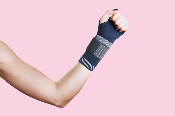 A white woman's arm is in a flexed position against a light pink background. She is wearing black nail polish and a black brace on her wrist, suggested that she's injured or experiencing chronic pain.