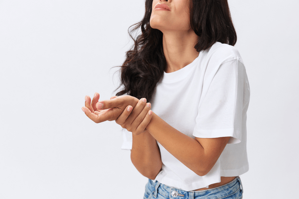 A woman with long, wavy brown hair and light brown skin is against a white background. She wears a cropped white T-shirt and jeans. She grimaces while holding one of her wrists, suggesting that she's experiencing chronic pain.