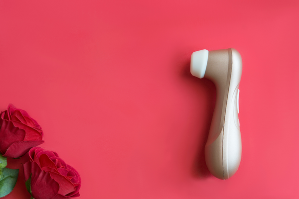 Against a red background, two roses are in the bottom left corner of the image, and a suction sex toy with a gold handle and a white mouth is on the right.