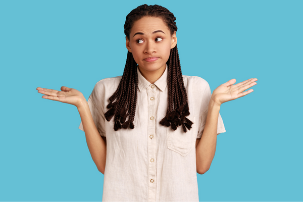 A Black woman with braids that go past her shoulders wears a white, button-up, short-sleeve shirt against a teal background. She opens her hands and shrugs.