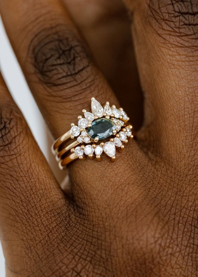 close up of a Black person's hand posing with a gorgeous gold engagement ring with a large sparkly green stone in the center and tons of tiny diamonds surrounding it