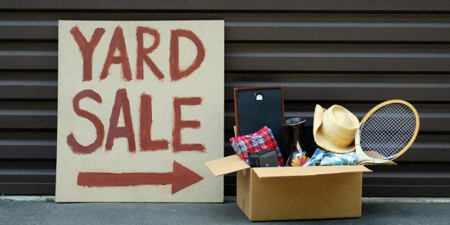 A Yard Sale sign hand written in red paint, set on the floor in front of a garage door. Next to it is a cardboard box filled to the brim with old items such as a cowboy hat and a tennis racket.