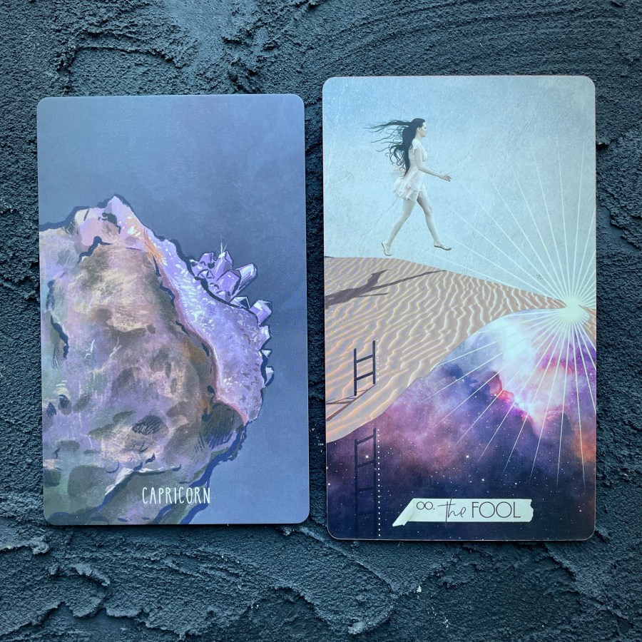Two cards in front of a navy blue background, right to left: Capricorn (the unclose surface of a precious gemstone) and the Fool (a woman Gallups across a beach)
