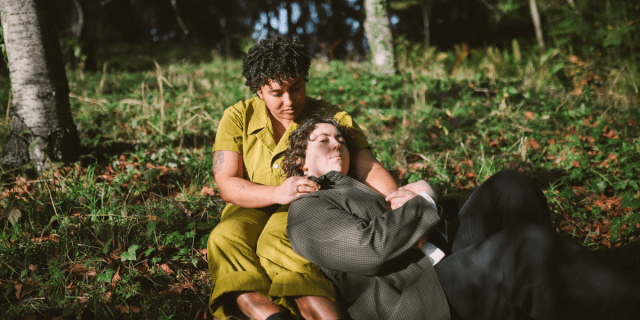 a couple sits outside in a field, looking worried and somber, holding each other