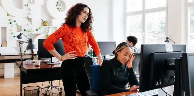 business manager overlooking a colleague struggling while working at a computer desk at the office.