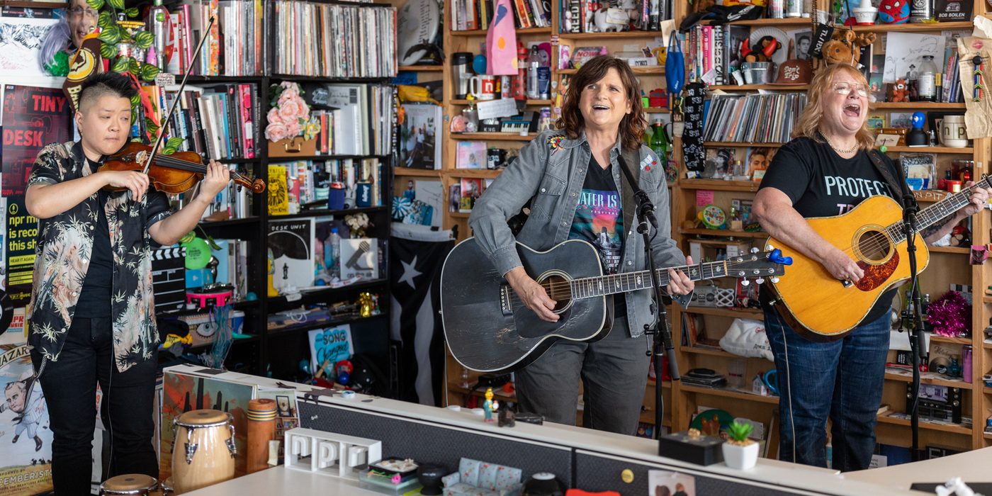 The Indigo Girls perform in a cramped library space with many books. They seem happy while singing.