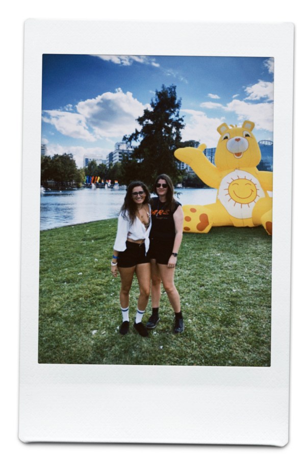 kayla and kristen stand in a field by a lake and there's a giant inflatable yellow care bear behind them
