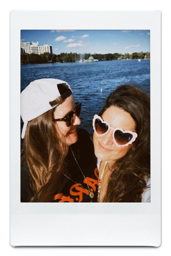 Kristen wears a backwards white ball cap and looks at Kayla who is wearing pink heart shaped sunglasses. there is a lake behind them
