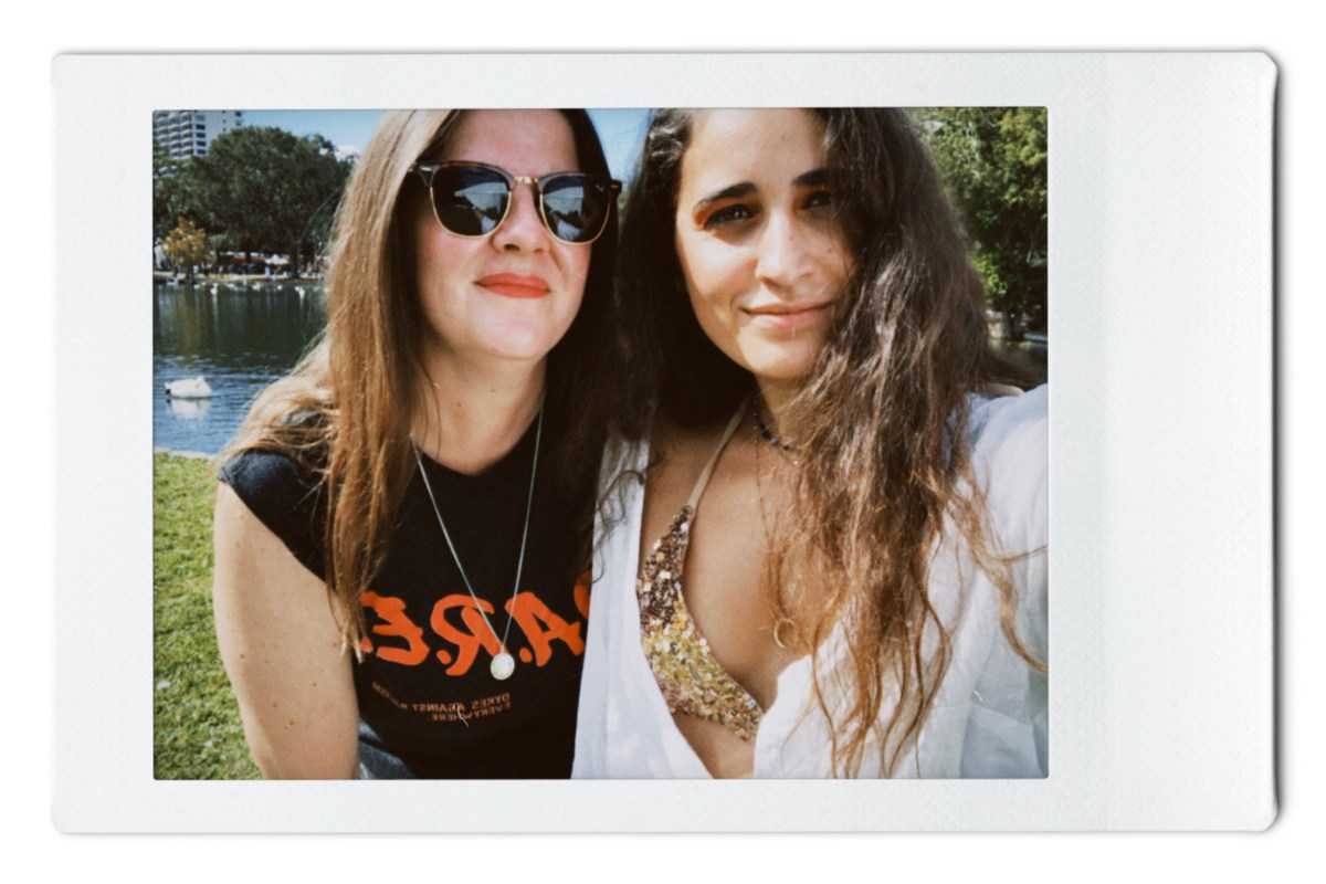 Kristen wears sunglasses and a shirt that says DARE (dykes against racism everywhere) and Kayla wears a gold bikini top with a white buttondown shirt