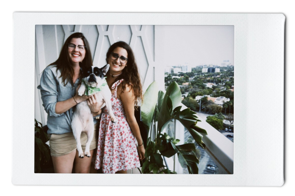 Kristen holds up Lola and Kayla is similing in a pink floral dress. They're on their balcony in Miami