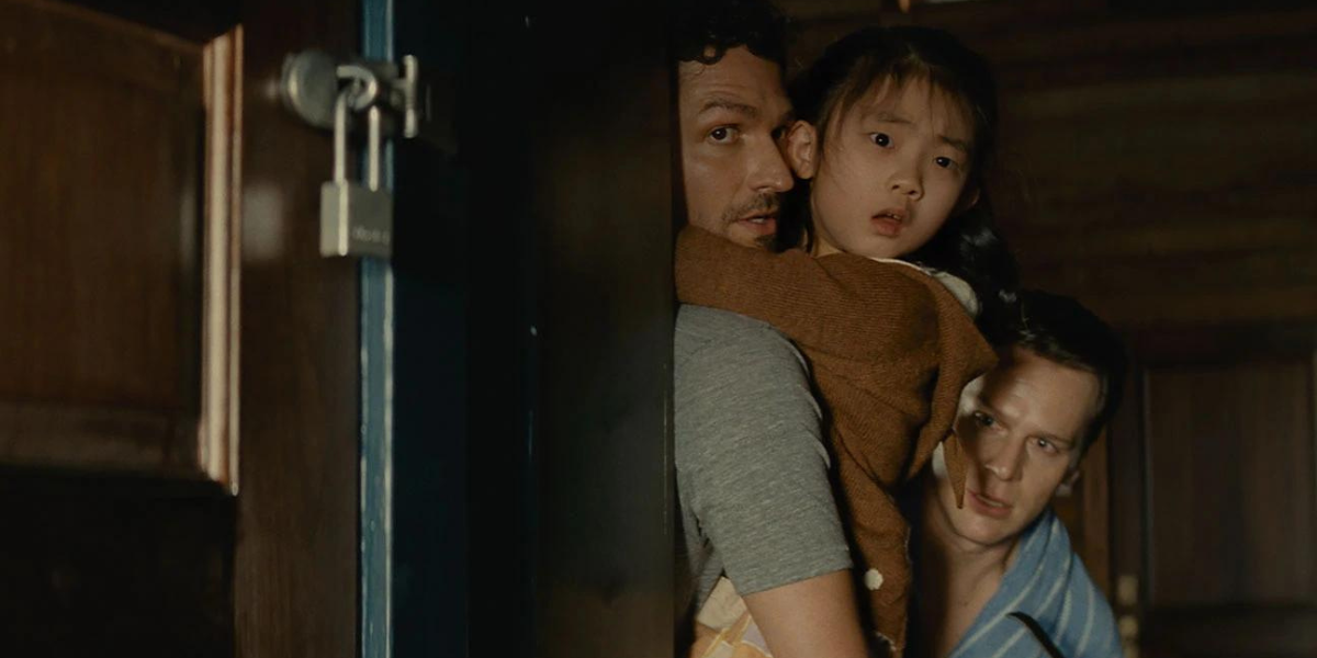 a still from Knock at the Cabin: two white men hold onto a young east Asian girl in the door of a cabin, looking afraid.