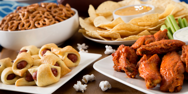 Super Bowl snack ideas: a bowl of pretzels, a plate of pigs in a blanket, a plate of wings and celery, and a plate of tortilla chips with a bowl of queso