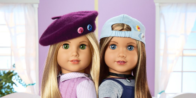 Two American Girl Dolls. One is blonde with green eyes and wears a purple beret and pink sweater vest over a striped buttondown. One is blonde with blue eyes in a backwards blue cap with a choker, a gray shirt, and a dark blue jumper over it.