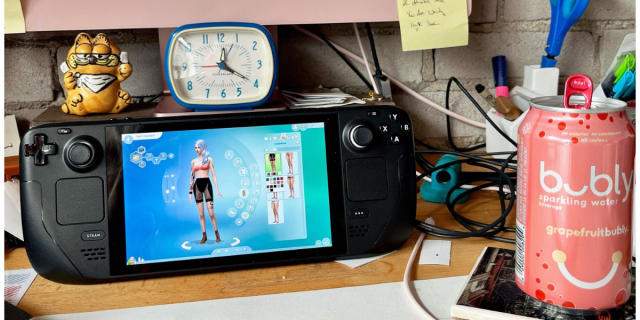 A Steam Deck playing the Sims sits on a desk with a Garfield figurine, an alarm clock, and a can of grapefruit Bubly.