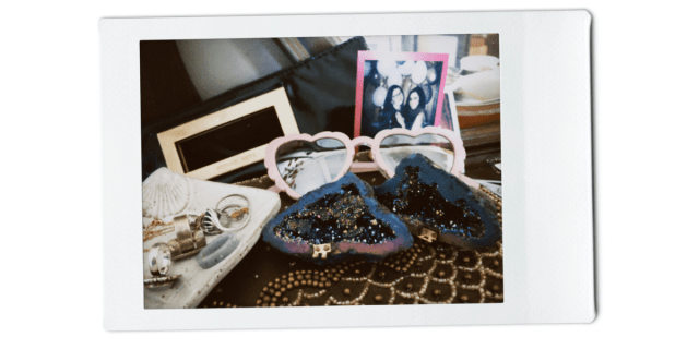 on a desk sits a beaded jewelry box, a black clutch purse, a gray ring tray with assorted rings on it, a polaroid of Kayla Kumari Upadhyaya and Kristen Arnett on new year's 2023, and a very cool rock, bisected in the middle and sparkly on the inside