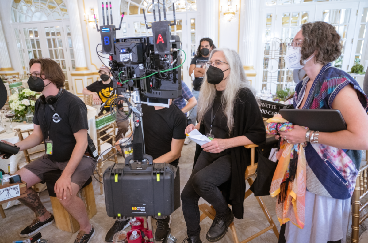 Annette Haywood-Carter, a white queer person with long grey hair and a Black Kn95 mask, sits in a director's chair working on set.
