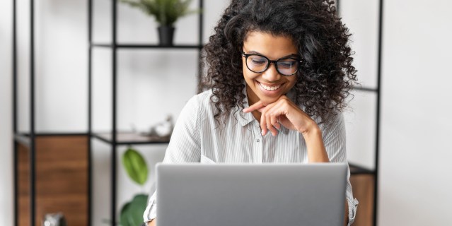 a young Black woman with glasses and curly hair sits at a desk smiling at her laptop, presumably communicating with someone she enjoys chatting with at work