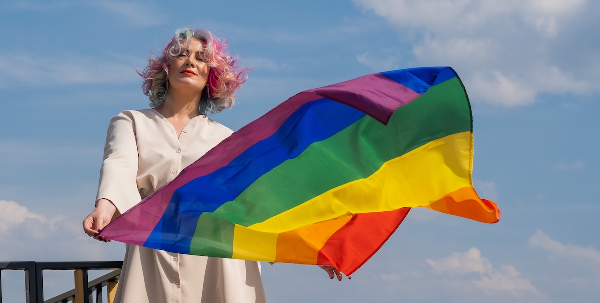 woman with pink short hair stands against a clear blue sky, holding a rainbow flag billowing in the wind, looking content and sure of herself