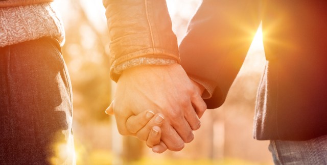close up of two friends holding hands. all we can see are the two hands, happily held together, with bright sunlight in the background.
