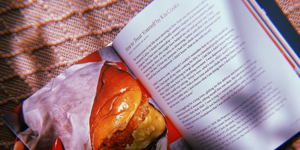 A photo of a popeyes chicken sandwich next to an essay about said sandwich inside of the book "Recipe for Disaster". A hand with painted pointy red nails holds the book open.
