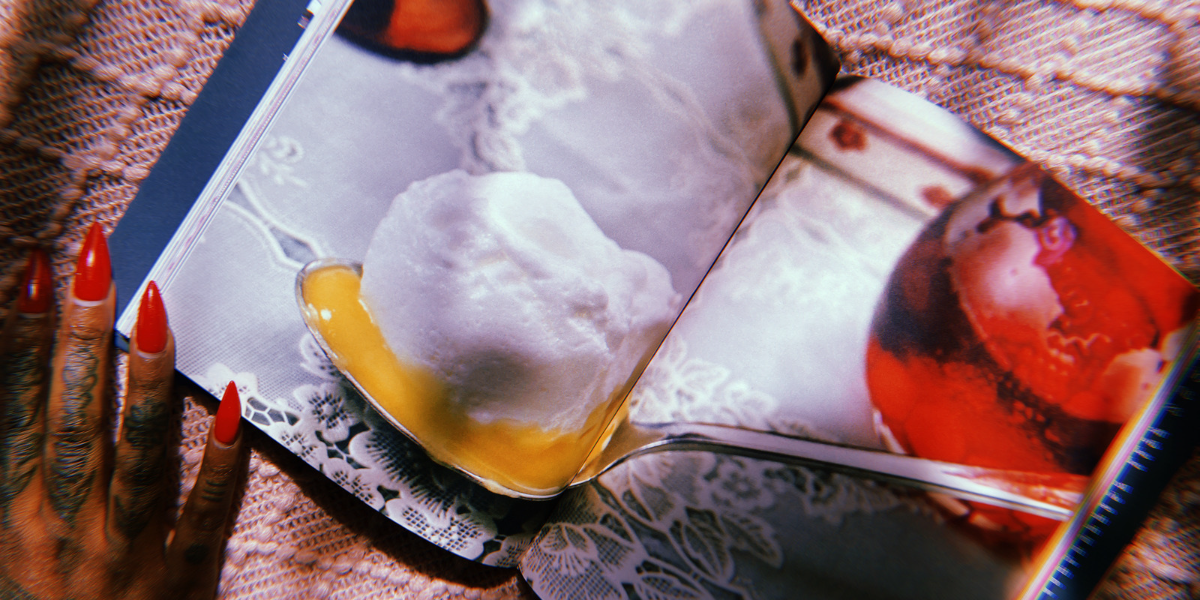 A photo of a spoon with cream on it inside the book "REcipe for disaster". A hand with painted pointy red nails holds the book open