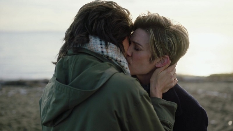 A Million Little Things: After Greta accepts her marriage proposal, she and Katherine share a kiss. Katherine has her back to the audience; she is wearing an olive green jacket with a hood and a multicolored scarf. Greta is wearing a dark blue pea coat.