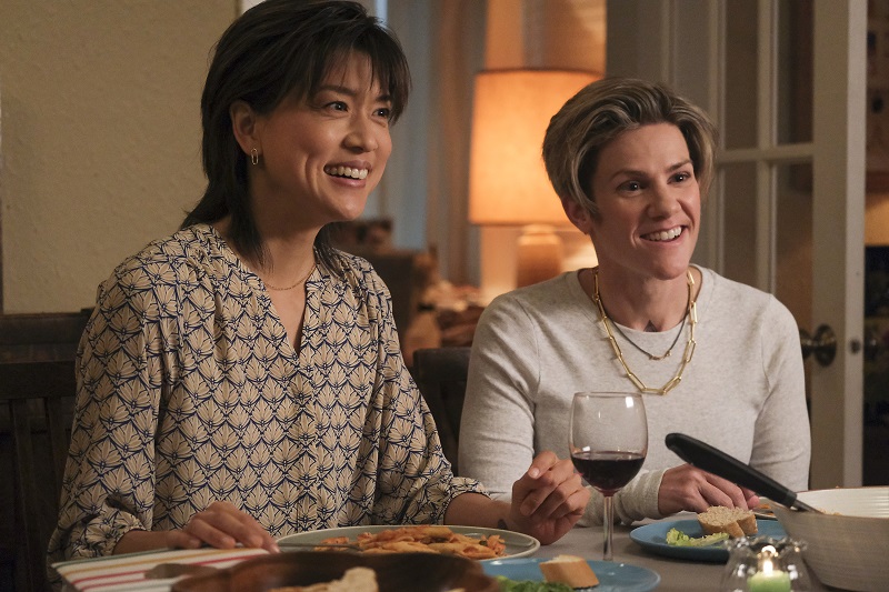 A Million Little Things: Katherine and Greta sit next to each other at Greta's parents' dinner table. Katherine is on the left, wearing a 3/4 sleeve floral blouse, and Greta is on the right, wearing a henley with a gold paperclip chain and small necklace.