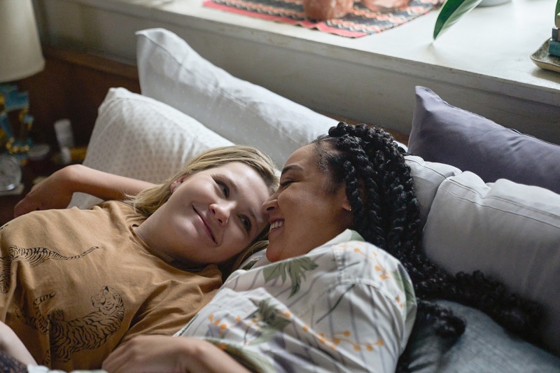 Accused: Esme and Aaliyah cuddle together on their bed. Esme's on the left, laying over Aaliyah's outstretched arm, and is beaming at her girlfriend. She's wearing a mustard graphic t-shirt with tigers on it. Aaliyah is on the right, smiling brightly at her girlfriend, while wearing a button up with stripes and palm trees.