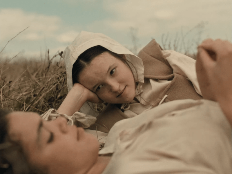 Bella Ramsey's Evelyn looks lovingly down at Mary, laying next to hear in a field