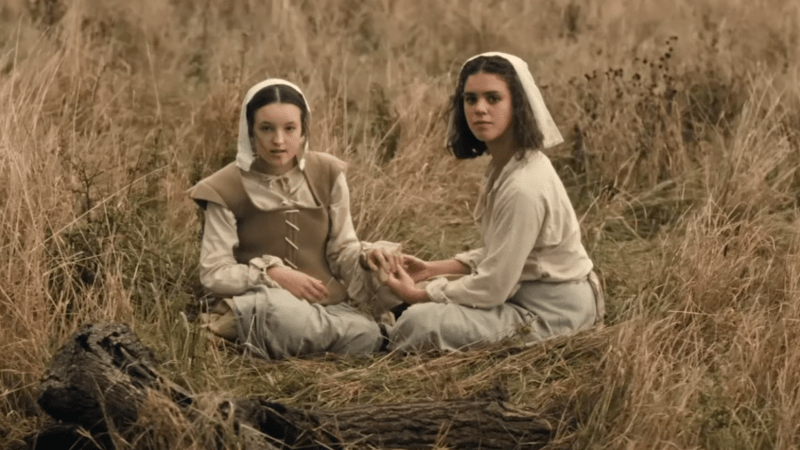 Requiem: Evelyn and Mary hold hands in a field, looking nervously at passing (off-screen) men