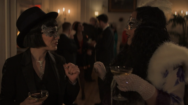 The Watchful Eye: Alex and Ginny in full masquerade garb smile at each other while holding fancy champagne 