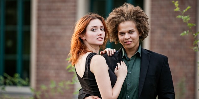 A Black butch with an afro holds their white femme partner with red hair by the waist, both are in formal wear and are staring at the camera.