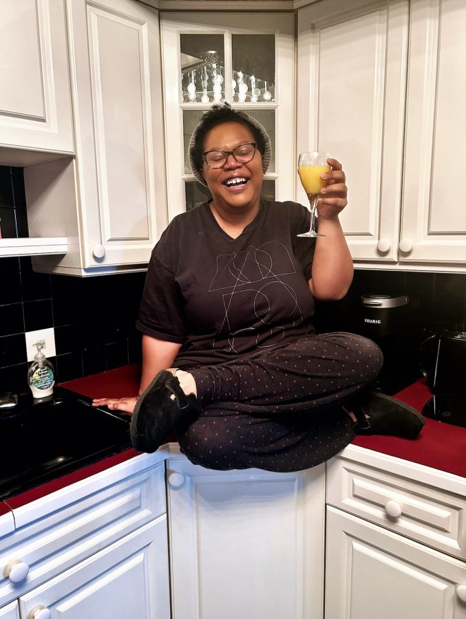 Carmen, a Black woman, sits on a kitchen counter, legs crossed, wearing a you do you tee, casual pants and birkenstocks. she is holding orange juice in a wine glass probably a mimosa and smiling