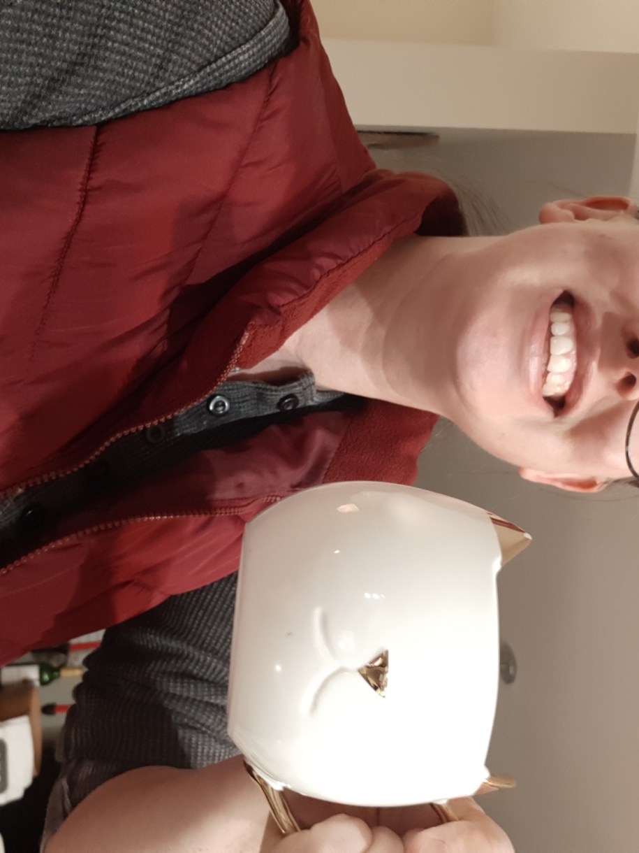 casey is a white woman with glasses who is holding a mug in the shape of a cat's face up to the camera - she is wearing glasses