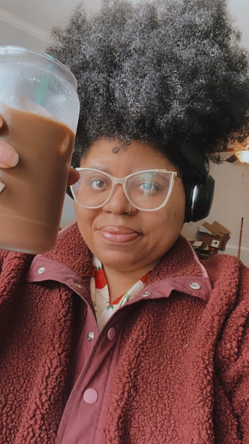 Dani, a black woman who here has a towering mass of curls on her head and is wearing glasses and a wine colored fleece is holding up a cup of iced coffee to the camera and smiling lightly