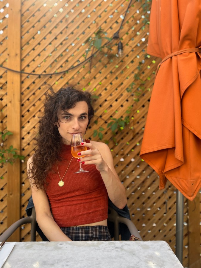 Drew a white woman with long curly hair is wearing a plaid skirt maybe and a rust colored sleeveless top. she outside sipping on either orange wine or an aperol spritz and looking at the camera