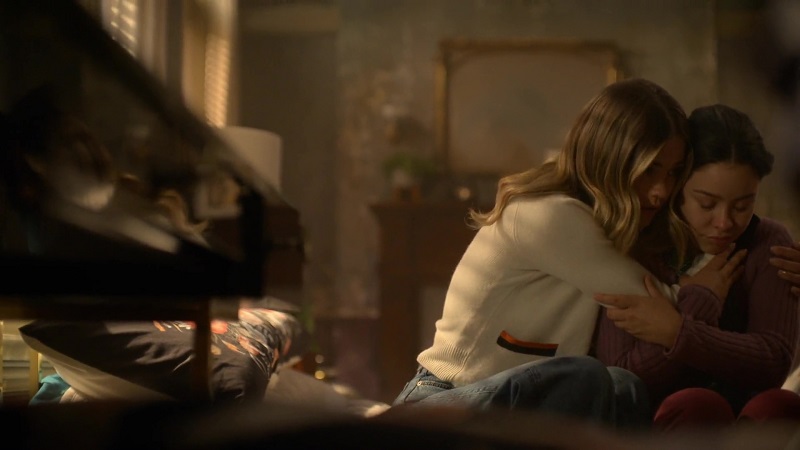 Good Trouble: Sitting on Mariana's bed, Callie wraps her sister in an embrace. Callie, on the left, is wearing a white sweater with a rainbow patch on the pocket. Her arms are wrapped around her sister and Mariana is holding on tight. Mariana is wearing a purple sweater and maroon pants. 