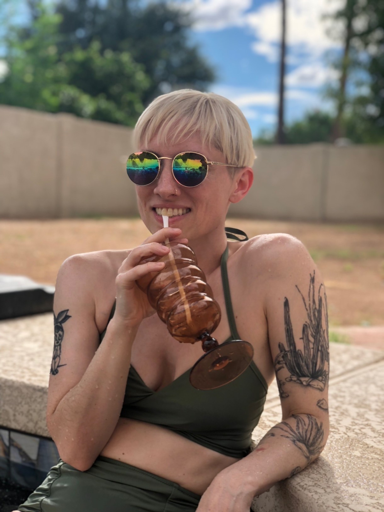 Laneia a white woman wears a green bathing suit and rainbow reflective round sunglasses and leans on the edge of a pool while sippping a beverage through a straw and smiling.