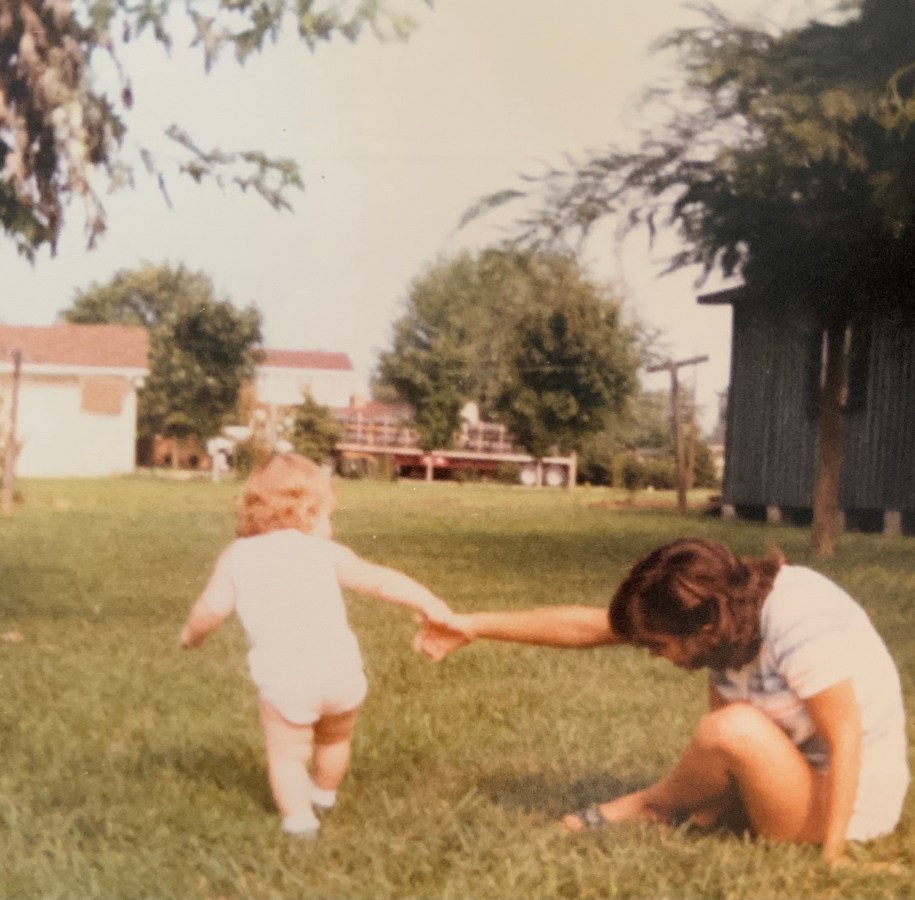 In a sunny photo from the early 80s, Laneia is a toddler with her back to the camera, wearing a white onesie and pulling her mother up from the grass so she'll follow her.