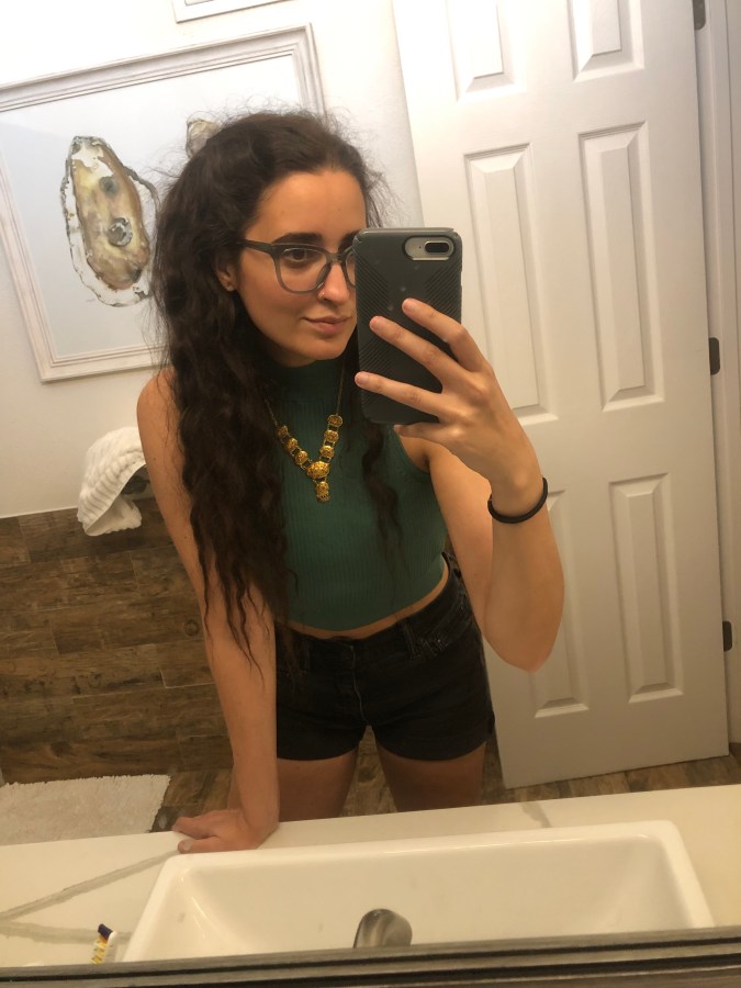 Kayla Kumari Upadhyaya wears a cropped green knit tank and a gold necklace over black jean shorts. Her hair is done in two small side partial-pigtails. She is wearing blue glasses.