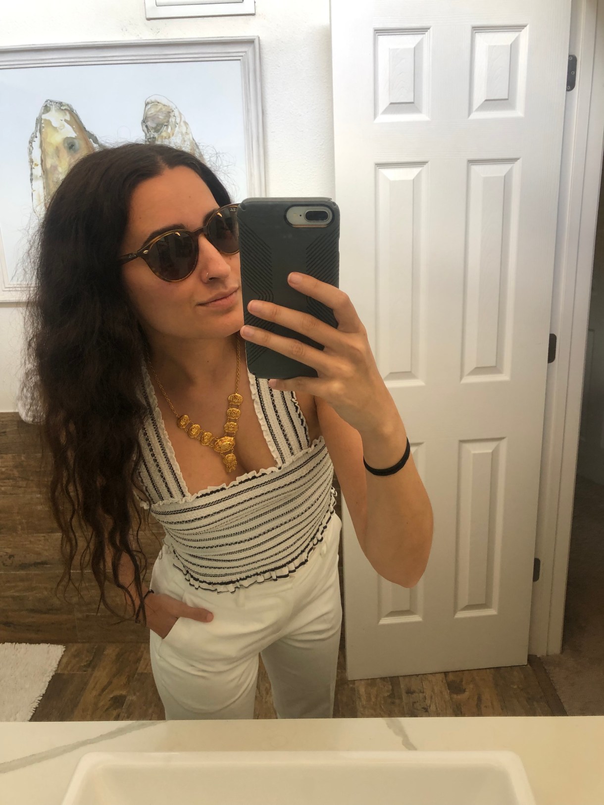 Kayla Kumari Upadhyaya is wearing sunglasses and a white an blue striped stretchy cropped tank with a gold necklace as well as tailored white pants. Her hair is down and parted in the middle.