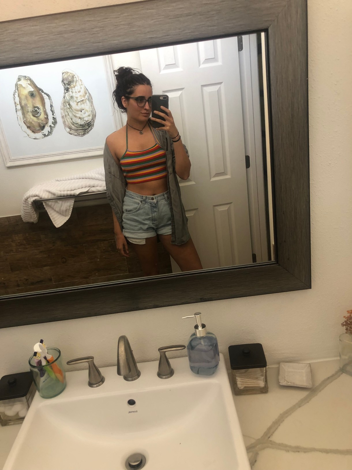 Kayla Kumari Upadhyaya wears a rainbow striped super cropped halter top over cut off lightwash jean shorts and also a gray buttondown that's open. She wears blue framed glasses, a brown choker, and her hair is wet and in a bun.