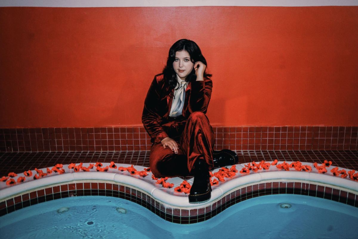 Lucy Dacus kneels by a heart shaped pool in a red velvet suit