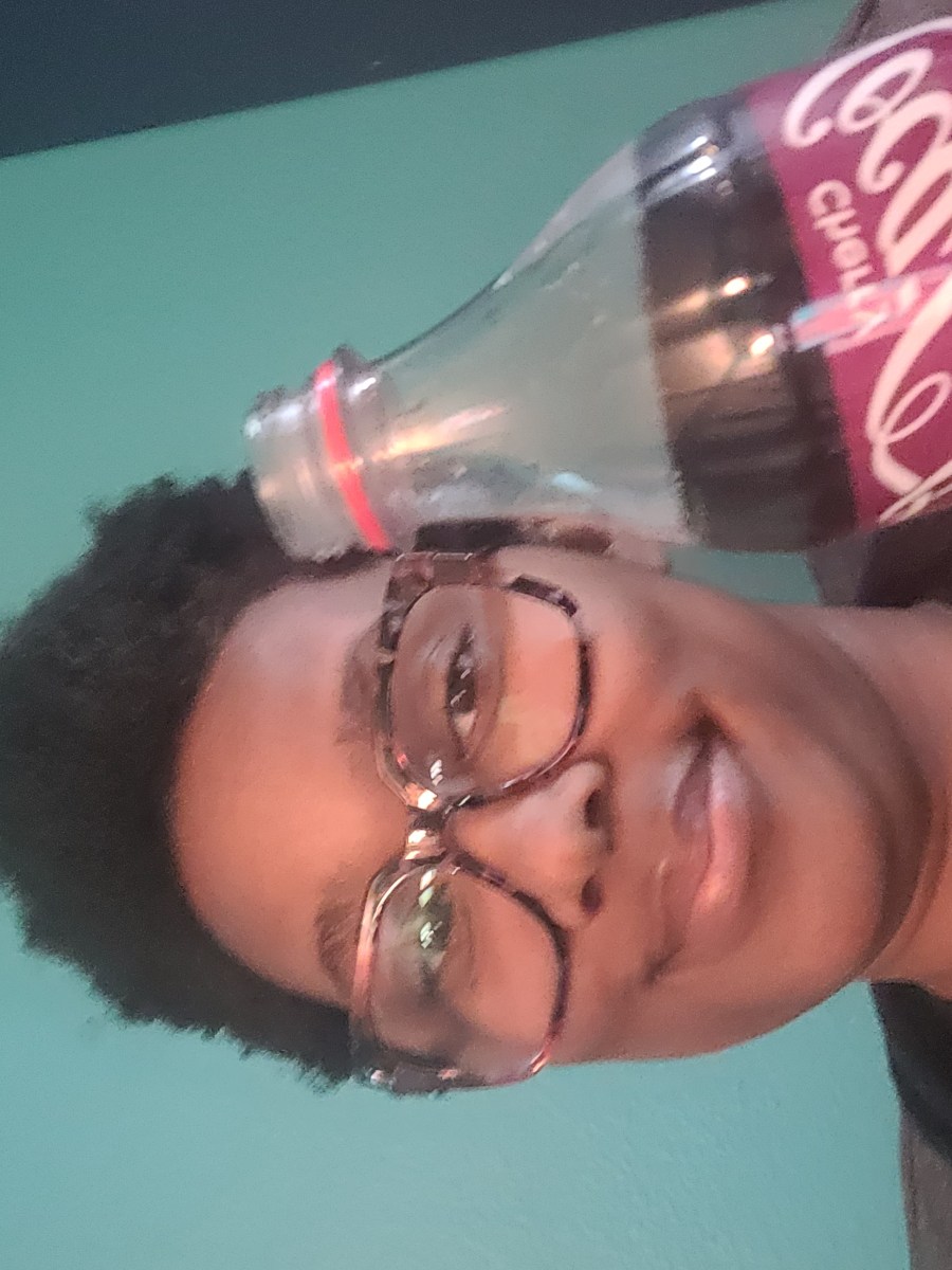 Sa'iyda, a Black woman wearing glasses holds up a cherry coke to the camera and smiles