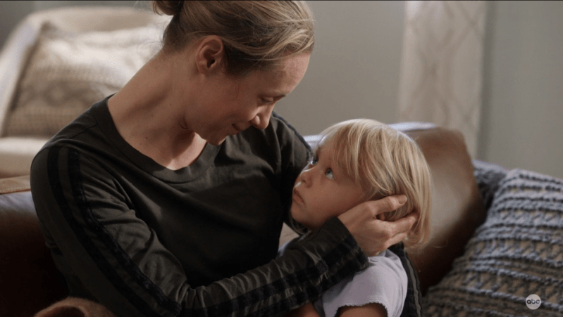 Maya, a blond woman with her hair in a bun, holds her three year old self in her arms (a blond child), on her couch, in Station 19
