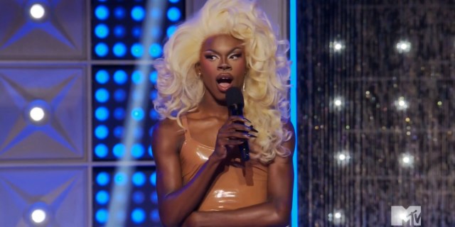Drag Race 1511 recap: Luxx Noir London holds a microphone with her arms crossed and looks shocked. She's in a tight burnt orange dress and a big blonde wig.