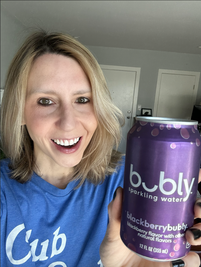 Riese is a white woman with shoulder length blonde hair who here is holding up a can of blackberry sparkling water and smiling