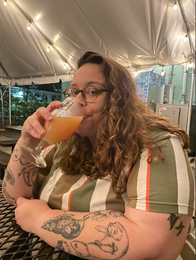 Stef, a white nonbinary person, drinks a beverage and looks at the camera. they are wearing a vertically striped top and have curly longer hair and glasses