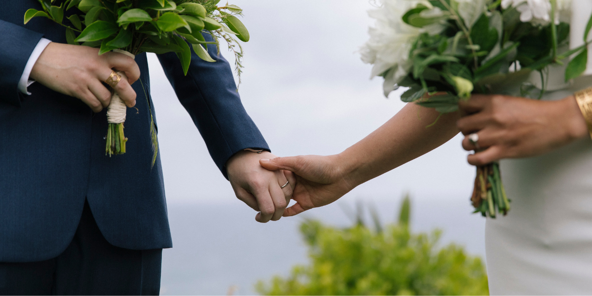 two people holding hands at an elopement in beautiful New Zealand, holding bouquets against moody blue skies and next to water